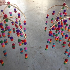 May include: Two wooden baby mobiles with colorful felt balls hanging from them. The mobiles are suspended from the ceiling and are made of natural wood.