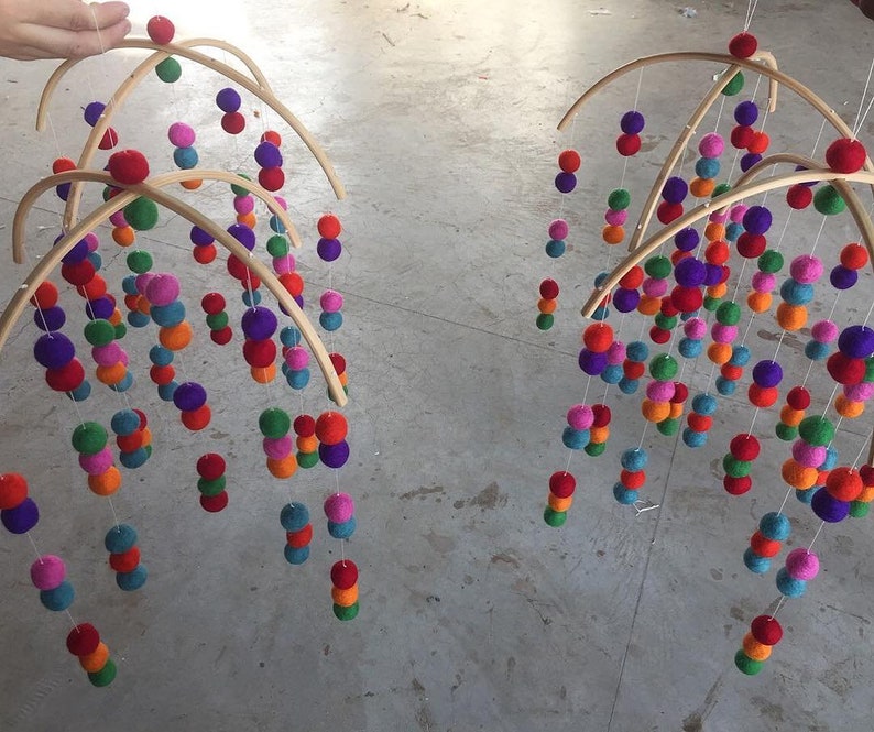 May include: Two wooden baby mobiles with colorful felt balls hanging from them. The mobiles are suspended from the ceiling and are made of natural wood.