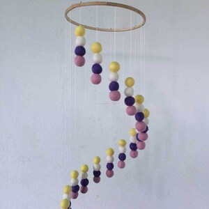 May include: A wooden hoop mobile with a spiral of felt balls in yellow, white, purple, and pink.