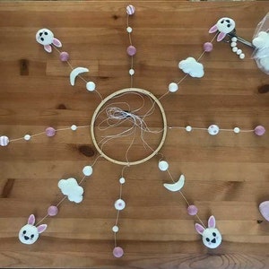 May include: A white wooden hoop with six strands of pink and white beads hanging from it. Each strand has a different felt ornament, including clouds, moons, and bunnies.