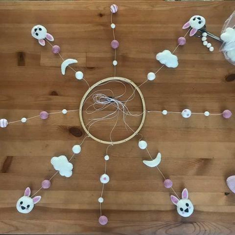 May include: A white wooden hoop with six strands of pink and white beads hanging from it. Each strand has a different felt ornament, including clouds, moons, and bunnies.