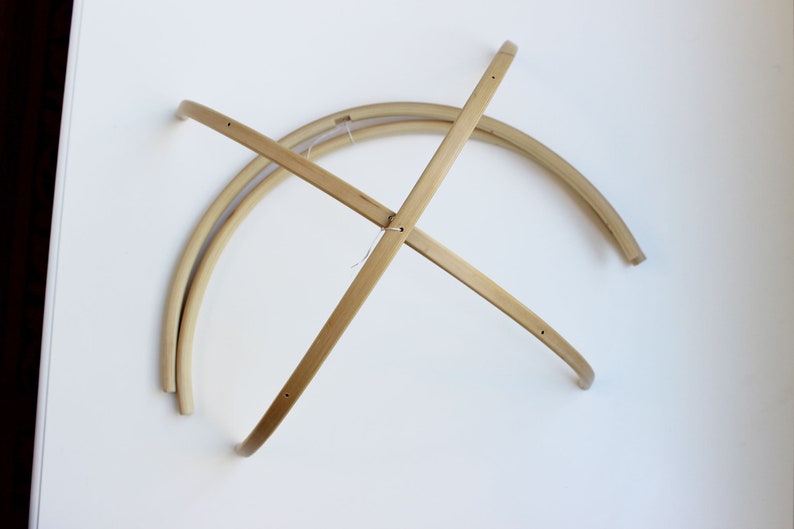 May include: A wooden mobile frame made of four curved pieces of wood that intersect in the center. The frame is designed to hold a mobile.