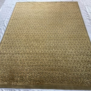 May include: A large, rectangular rug with a repeating geometric pattern in shades of tan and olive green. The rug's design features interlocking shapes, creating a textured surface. The rug has a border in a darker shade of green.