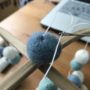 May include: A blue felt ball is being tied to a wooden mobile with white string. The mobile is made of two wooden dowels and has other felt balls in various colors, including white, light blue, and green.