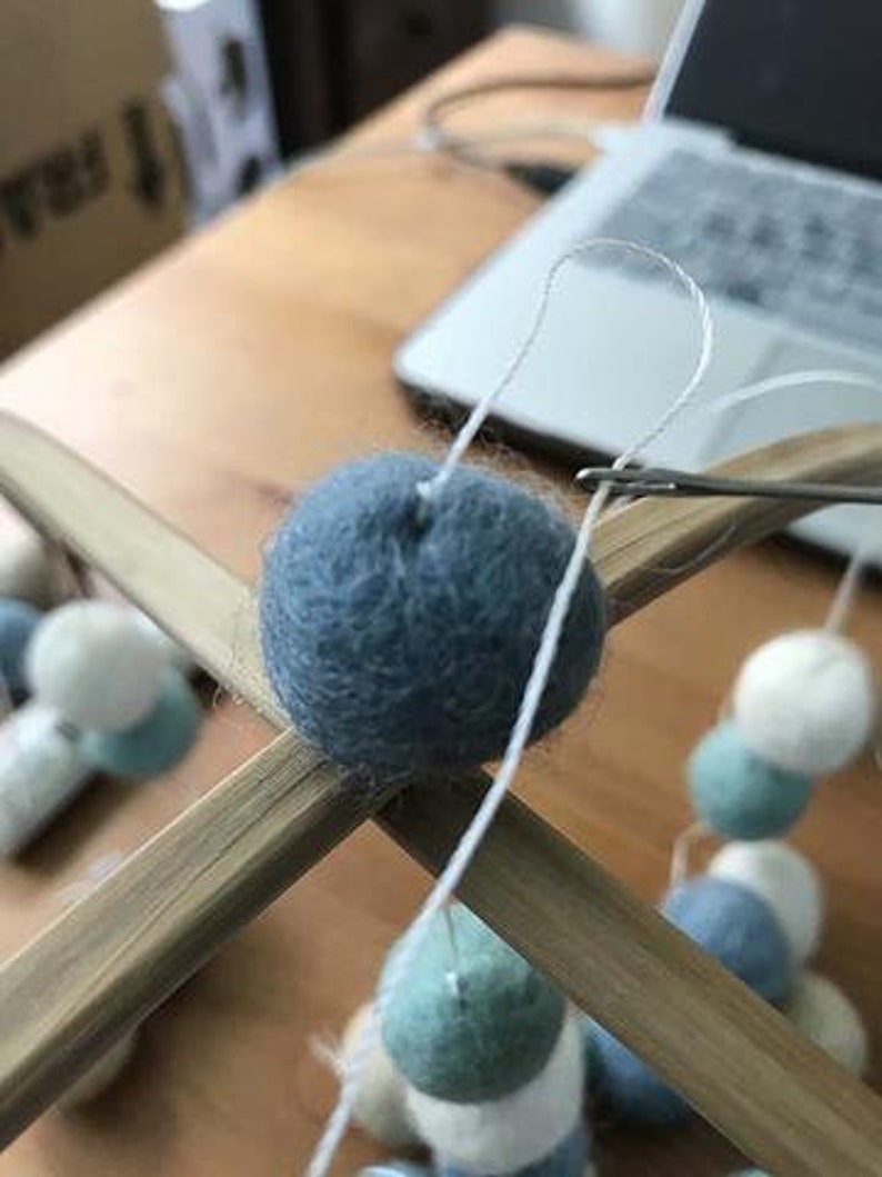 May include: A blue felt ball is being tied to a wooden mobile with white string. The mobile is made of two wooden dowels and has other felt balls in various colors, including white, light blue, and green.