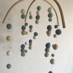 May include: A wooden mobile with multiple felt balls in shades of blue, green, and beige. The balls are suspended from a wooden frame and hang at different lengths.