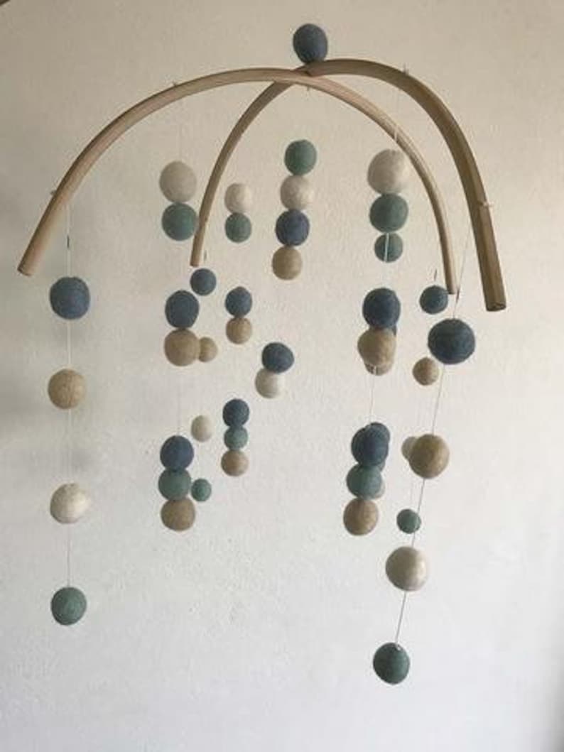 May include: A wooden mobile with multiple felt balls in shades of blue, green, and beige. The balls are suspended from a wooden frame and hang at different lengths.