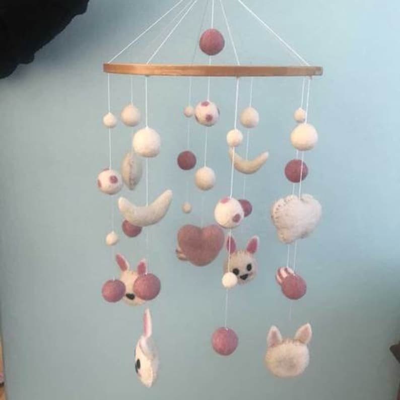 May include: A white and pink felt baby mobile with a wooden ring. The mobile features a variety of shapes, including stars, moons, hearts, and animals. The mobile is suspended from the ceiling by white strings.