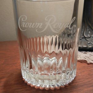 May include: Clear glass tumbler with the etched text "Crown Royal". The glass has a ribbed design on the lower portion and a rounded base. The glass is sitting on a wooden surface.