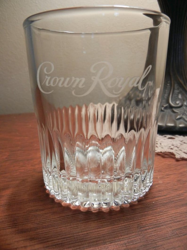 May include: Clear glass tumbler with the etched text "Crown Royal". The glass has a ribbed design on the lower portion and a rounded base. The glass is sitting on a wooden surface.