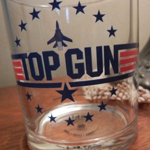 May include: Clear glass tumbler featuring the text "TOP GUN" in navy blue and red, with a navy blue jet graphic and star accents. The glass has a slightly flared shape and is designed for beverages.
