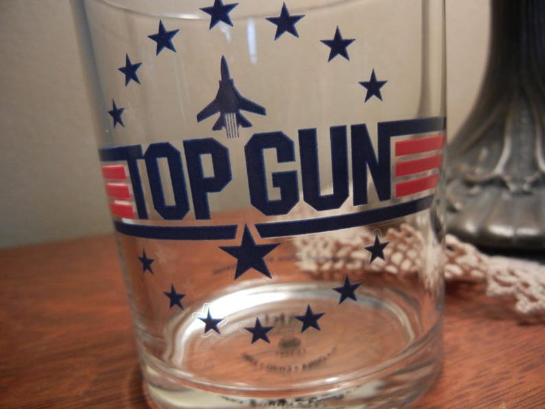 May include: Clear glass tumbler featuring the text "TOP GUN" in navy blue and red, with a navy blue jet graphic and star accents. The glass has a slightly flared shape and is designed for beverages.