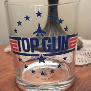 May include: A clear glass tumbler featuring the text "TOP GUN" in navy blue with red and white stripes. The design includes navy blue stars and a silhouette of a jet. The glass is cylindrical and appears to be empty.