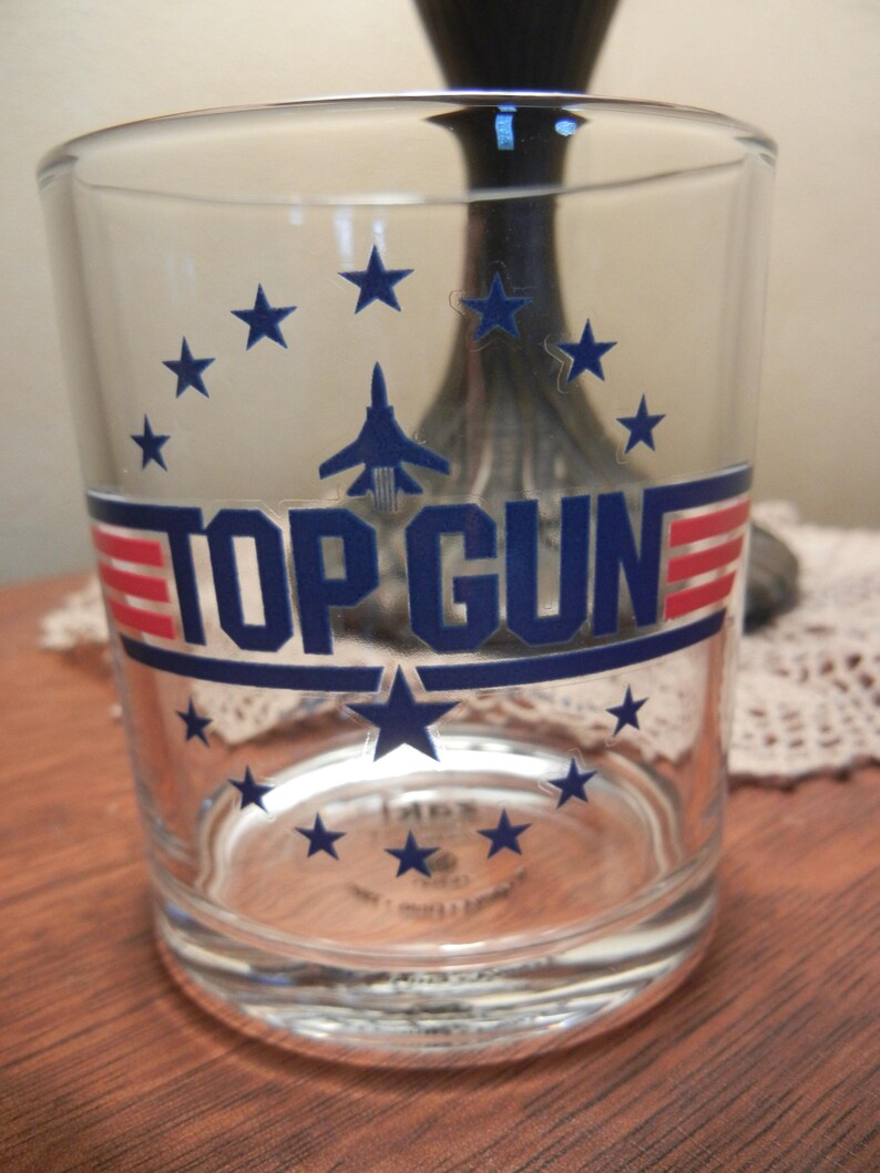 May include: A clear glass tumbler featuring the text "TOP GUN" in navy blue with red and white stripes. The design includes navy blue stars and a silhouette of a jet. The glass is cylindrical and appears to be empty.