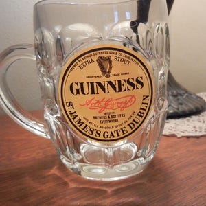 May include: A clear glass beer mug with a handle. The mug features a circular label with the Guinness logo, including the harp symbol and the words "Guinness Extra Stout". The label also includes the text "St. James's Gate Dublin".