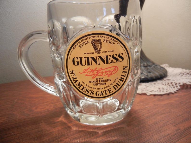 May include: A clear glass beer mug with a handle. The mug features a circular label with the Guinness logo, including the harp symbol and the words "Guinness Extra Stout". The label also includes the text "St. James's Gate Dublin".