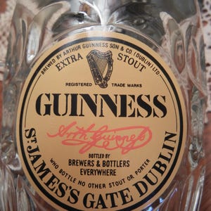 May include: A clear glass Guinness pint glass with a textured surface. The glass features a circular label with the Guinness logo, harp, and text including "St. James's Gate Dublin".
