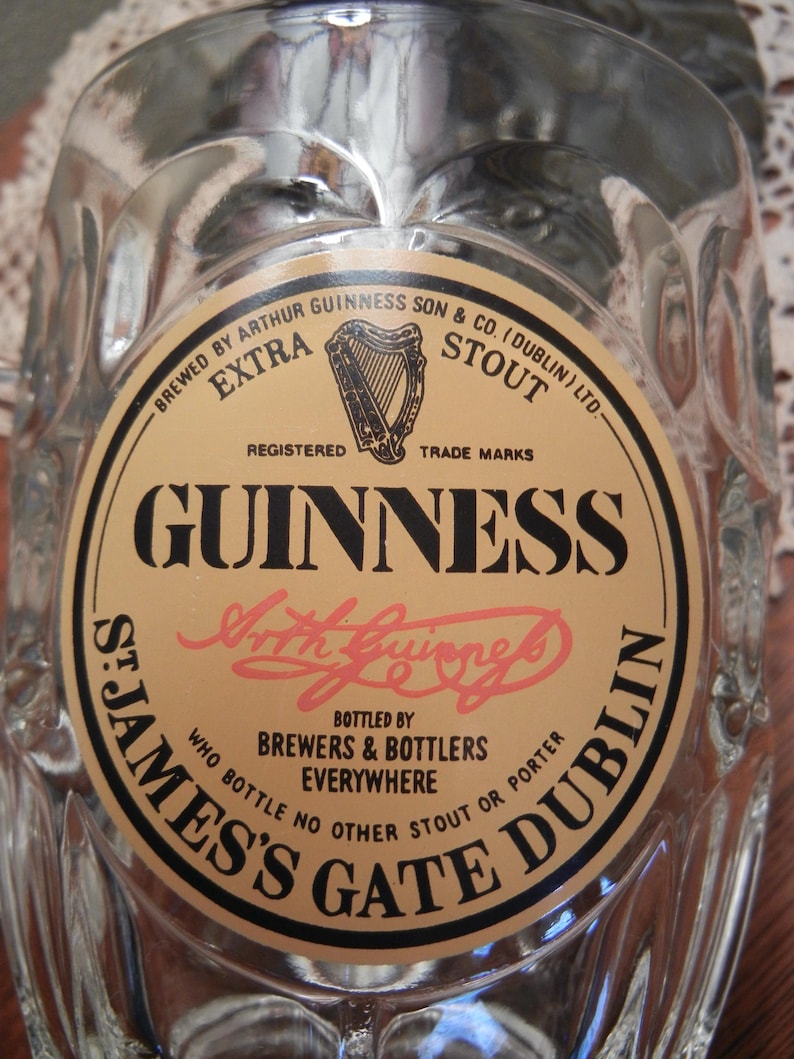 May include: A clear glass Guinness pint glass with a textured surface. The glass features a circular label with the Guinness logo, harp, and text including "St. James's Gate Dublin".