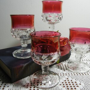 May include: Four vintage wine glasses with ruby red bowls and clear, patterned bases. The glasses are arranged on a dark blue book and a white lace tablecloth, with a rose in the background. The glasses have a classic, elegant design.