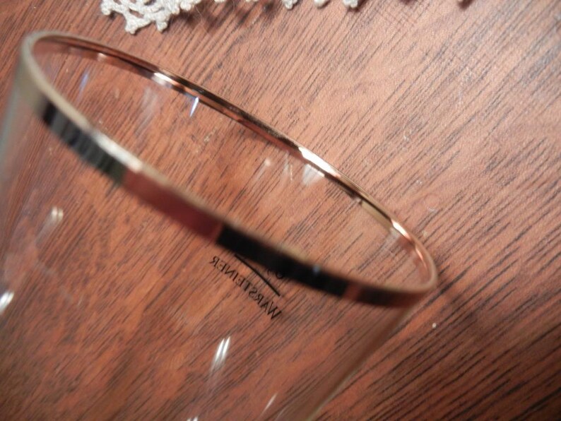 May include: A clear glass with a gold rim and the word "WARSTEINER" printed on the side. The glass is set against a brown wooden surface.