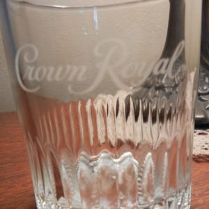 May include: A clear, textured glass with the words "Crown Royal" etched on the front. The glass has vertical ridges and a thick base. The glass is sitting on a wooden surface.