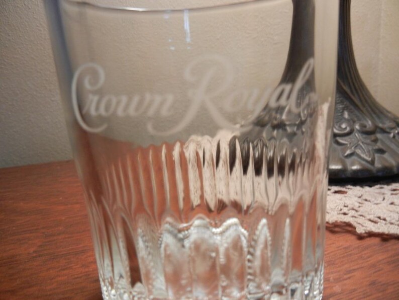 May include: A clear, textured glass with the words "Crown Royal" etched on the front. The glass has vertical ridges and a thick base. The glass is sitting on a wooden surface.