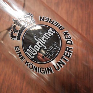 May include: Clear glass beer glass with the words "Warsteiner Premium Verum" and a crown logo. The text "Eine K&ouml;nigin Unter Den Bieren" is also printed on the glass. The glass is angled, showing the interior.