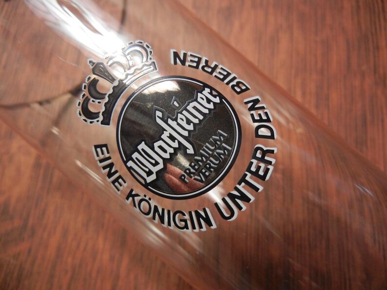 May include: Clear glass beer glass with the words "Warsteiner Premium Verum" and a crown logo. The text "Eine K&ouml;nigin Unter Den Bieren" is also printed on the glass. The glass is angled, showing the interior.