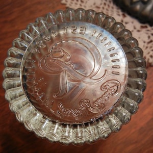 May include: A silver-colored, fluted baking mold with a decorative embossed design in the center. The center design features an ornate letter "R" and surrounding text. The mold is circular and sits on a wooden surface.