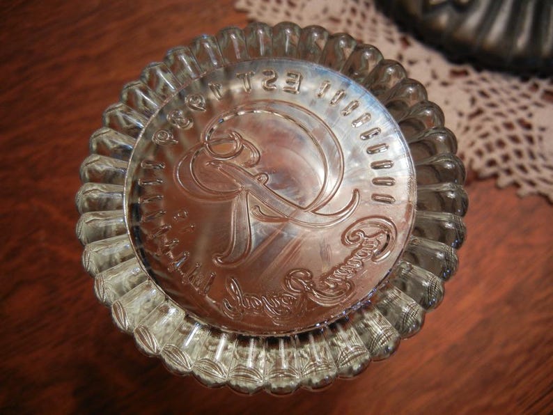 May include: A silver-colored, fluted baking mold with a decorative embossed design in the center. The center design features an ornate letter "R" and surrounding text. The mold is circular and sits on a wooden surface.