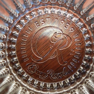 May include: Close-up of a clear glass with a textured, radial design. The center features a brown, circular emblem with the words "EST 1939" and "Crown Royal" in a stylized font. The glass has a reflective quality.
