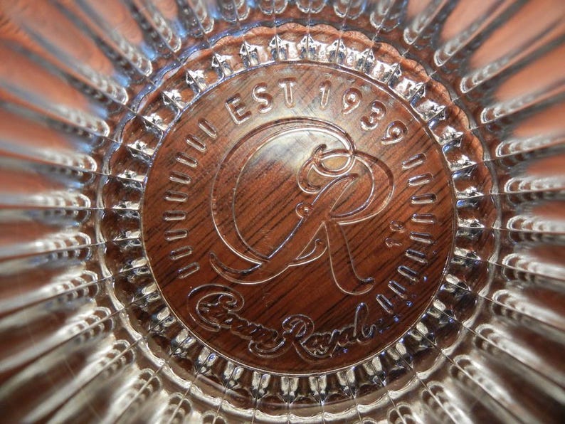 May include: Close-up of a clear glass with a textured, radial design. The center features a brown, circular emblem with the words "EST 1939" and "Crown Royal" in a stylized font. The glass has a reflective quality.