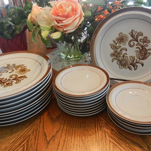 Puede incluir: Un conjunto de vajilla de cerámica vintage, que incluye platos y platillos. Los platos presentan un diseño floral en marrón y azul, con un borde marrón. Los platillos y platos están apilados sobre una superficie de madera.