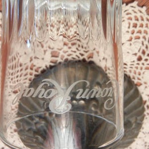 May include: Clear glass tumbler with vertical ribbed design. The glass has the words "Crown Royal" etched on the side. The glass is set on a decorative metal base, with a white lace doily in the background.