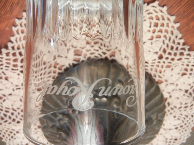 May include: Clear glass tumbler with vertical ribbed design. The glass has the words "Crown Royal" etched on the side. The glass is set on a decorative metal base, with a white lace doily in the background.