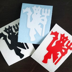May include: Three vinyl stickers featuring a stylized devil with a trident. One sticker is black, one is white, and one is red. The stickers are on a white backing, and the background is a dark brown surface.