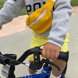 K&ouml;nnte beinhalten: Ein Kind tr&auml;gt ein graues Sweatshirt mit dem Wort "Polo" darauf, gelbe Shorts und eine gelbe Bauchtasche mit einem wei&szlig;en Rei&szlig;verschluss und f&auml;hrt ein blaues Fahrrad mit schwarzem Lenker.