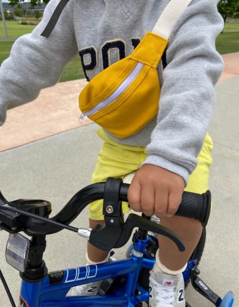 K&ouml;nnte beinhalten: Ein Kind tr&auml;gt ein graues Sweatshirt mit dem Wort "Polo" darauf, gelbe Shorts und eine gelbe Bauchtasche mit einem wei&szlig;en Rei&szlig;verschluss und f&auml;hrt ein blaues Fahrrad mit schwarzem Lenker.