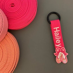 May include: Pink personalized keychain with the name Hailey embroidered in white. The keychain features a pink ballerina shoe charm with sequins.