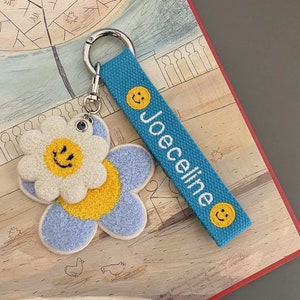 May include: A blue and white fuzzy flower keychain with a smiling face. The keychain is attached to a blue strap with the name "Joceline" embroidered on it.