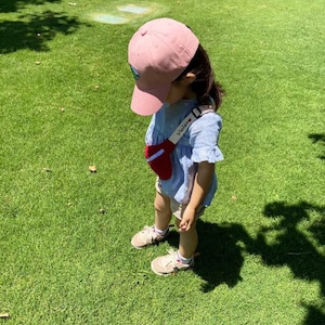 K&ouml;nnte beinhalten: Ein kleines Kind tr&auml;gt eine rosafarbene Baseballkappe mit einem gr&uuml;nen Logo, ein blau-wei&szlig; gemustertes Hemd und eine rote Bauchtasche mit dem aufgestickten Wort "Violet". Das Kind steht auf einem gr&uuml;nen Grasfeld.