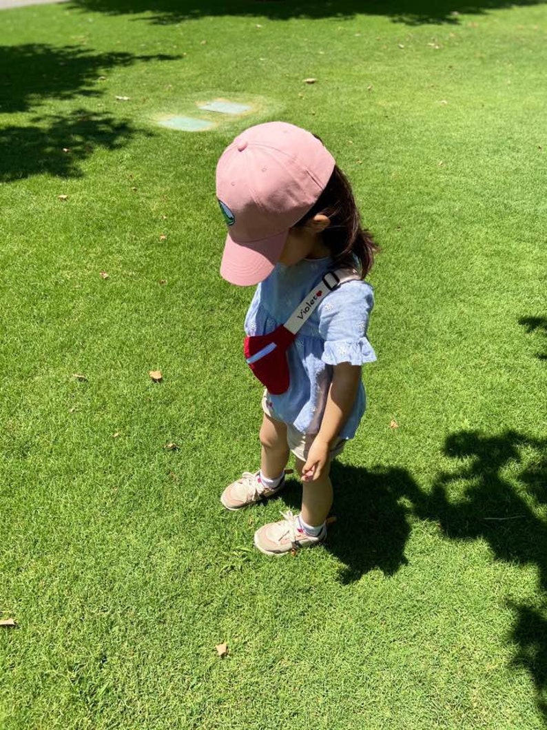 K&ouml;nnte beinhalten: Ein kleines Kind tr&auml;gt eine rosafarbene Baseballkappe mit einem gr&uuml;nen Logo, ein blau-wei&szlig; gemustertes Hemd und eine rote Bauchtasche mit dem aufgestickten Wort "Violet". Das Kind steht auf einem gr&uuml;nen Grasfeld.