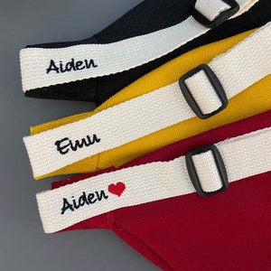 K&ouml;nnte beinhalten: Drei Stofftaschen mit wei&szlig;en Tr&auml;gern und schwarzen Schnallen. Die Tr&auml;ger sind mit den Namen "Aiden", "Euu" und "Aiden" mit einem roten Herzen personalisiert.