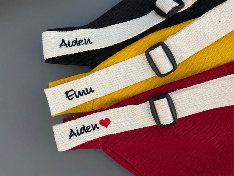 K&ouml;nnte beinhalten: Drei Stofftaschen mit wei&szlig;en Tr&auml;gern und schwarzen Schnallen. Die Tr&auml;ger sind mit den Namen "Aiden", "Euu" und "Aiden" mit einem roten Herzen personalisiert.