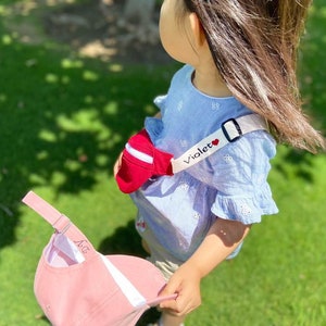 K&ouml;nnte beinhalten: Ein junges M&auml;dchen tr&auml;gt ein blau-wei&szlig;es Blumenkleid mit einem wei&szlig;en Band und einer rot-wei&szlig;en Tasche. Das Band ist mit dem Wort "Violet" und einem roten Herzen bestickt. Das M&auml;dchen h&auml;lt eine rosafarbene Baseballkappe mit dem Wort "Papa" darauf.
