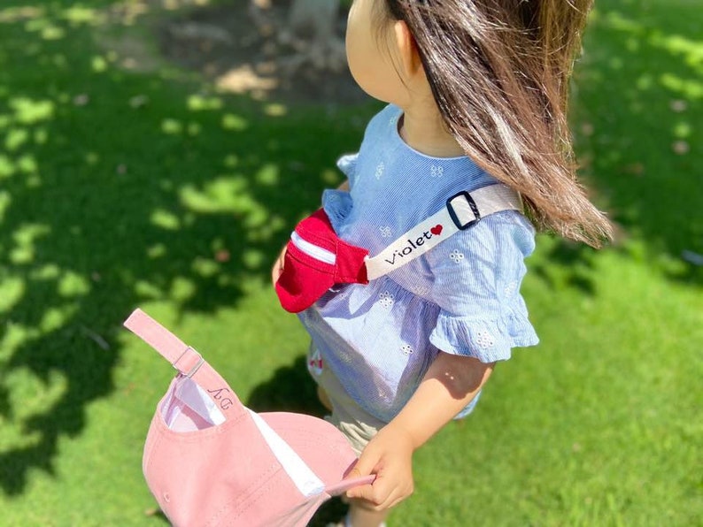 K&ouml;nnte beinhalten: Ein junges M&auml;dchen tr&auml;gt ein blau-wei&szlig;es Blumenkleid mit einem wei&szlig;en Band und einer rot-wei&szlig;en Tasche. Das Band ist mit dem Wort "Violet" und einem roten Herzen bestickt. Das M&auml;dchen h&auml;lt eine rosafarbene Baseballkappe mit dem Wort "Papa" darauf.