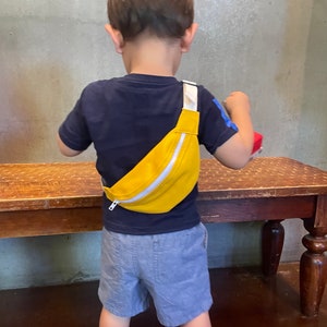 K&ouml;nnte beinhalten: Ein kleines Kind tr&auml;gt ein blaues Shirt und graue Shorts und eine gelbe Bauchtasche mit einem wei&szlig;en Rei&szlig;verschluss. Die Bauchtasche wird &uuml;ber dem K&ouml;rper getragen.