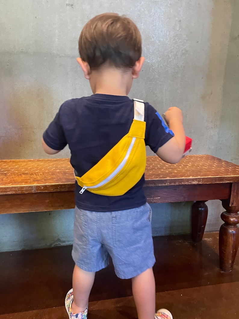 K&ouml;nnte beinhalten: Ein kleines Kind tr&auml;gt ein blaues Shirt und graue Shorts und eine gelbe Bauchtasche mit einem wei&szlig;en Rei&szlig;verschluss. Die Bauchtasche wird &uuml;ber dem K&ouml;rper getragen.