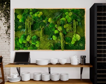May include: A green moss wall art piece with a wooden frame, hanging on a white wall above a wooden shelf with white dishes and a black laptop.