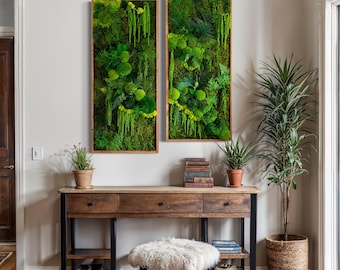 May include: Two framed moss wall art pieces with a wooden frame, hanging on a white wall above a wooden console table with two drawers and a lower shelf. The table has a white faux fur stool in front of it. A potted plant sits on the table and another potted plant sits on the floor next to the table.